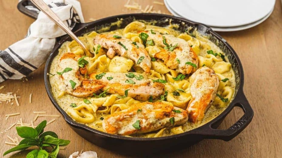 A cast iron skillet with creamy, cheesy tortellini pasta and chicken tenderloin in a pesto sauce.