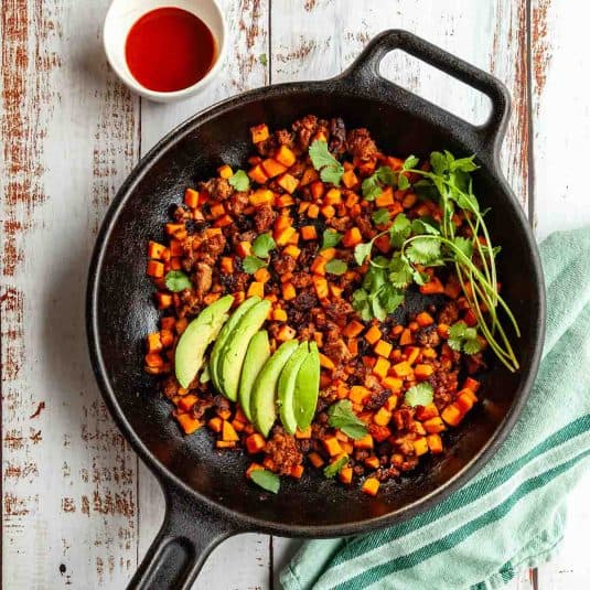 A cast iron skillet filled with chorizo and sweet potato hash with sliced avocado and cilantro on top.