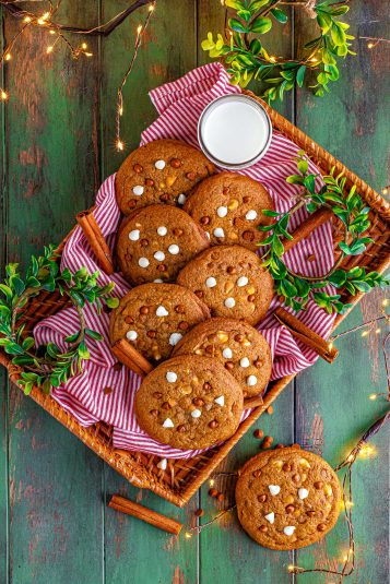 Close up side view of a decorative wooden tray filled with gingerbread chocolate chip cookies, stacked in a way so they're leaning back and showing off a good view to the camera, pictured with cinnamon sticks, holly, and decorative twinkle lights.