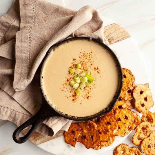 A skillet filled with Guinness beer cheese dip, topped with chopped green onions, and cayenne, with pretzel crackers on the side.