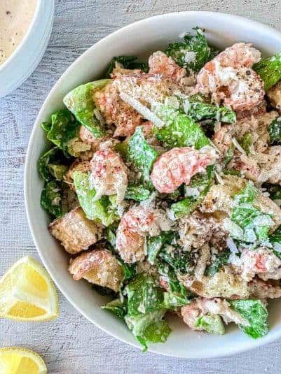 A fresh Lobster Caesar salad in a white bowl, featuring romaine lettuce, croutons, and grated Parmesan cheese, with a creamy dressing.