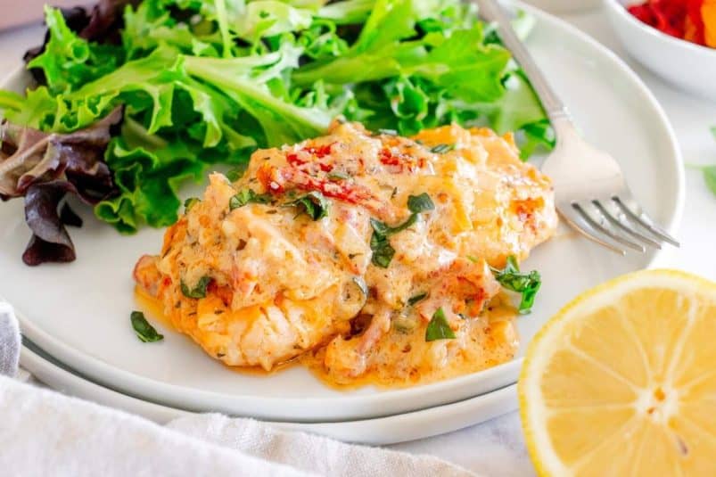 A plate of creamy Tuscan marry me salmon served with a side of fresh green salad and half a lemon, with a fork placed on the plate.