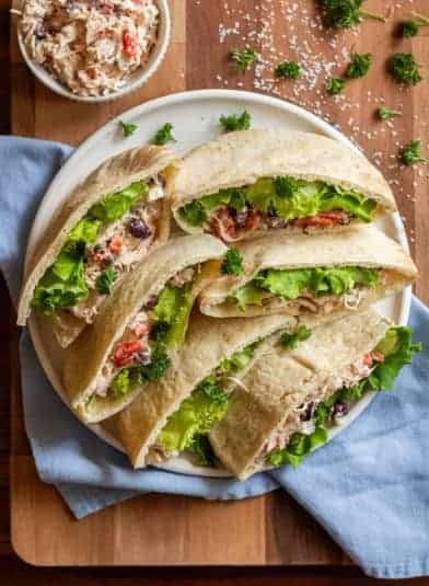 Chicken salad pitas are layered on a round, white plate. The plate is sitting on a cutting board and a blue napkin. There is fresh parsley garnishing the plate and a small bowl of chicken salad in the top left corner.