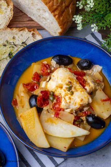 A blue bowl of tender white fish in a stew of potatoes, cheery tomatoes and black olives on a table with sliced bread.