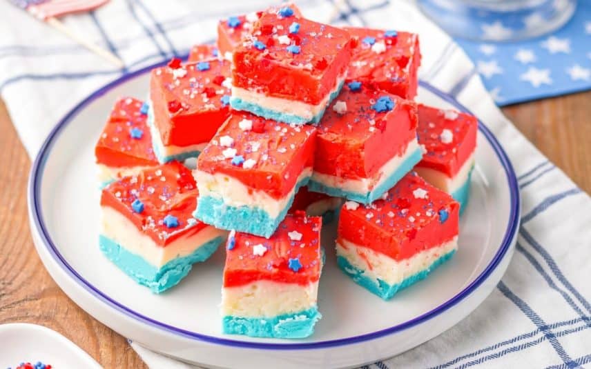 A plate of Patriotic red white and blue fudge squares topped with star-shaped sprinkles, arranged on a white plate with a blue rim on a checkered cloth.