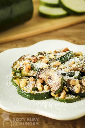 Side shot of sauteed zucchini and walnuts with parmesan cheese on a white decorative plate with raw zucchini on a cutting board in the background.