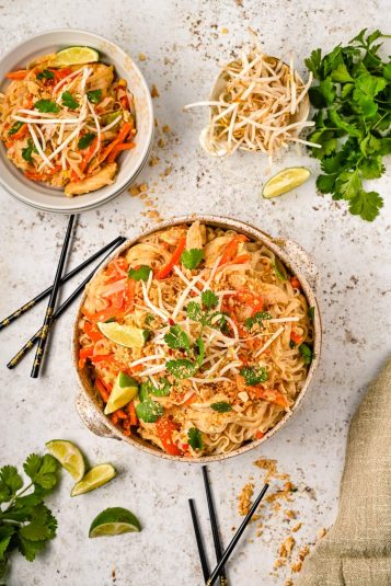 Overhead view of two bowls of easy chicken pad Thai garnished with bean sprouts, lime wedges, and cilantro, accompanied by chopsticks and scattered herbs and lime slices on a light-colored surface.