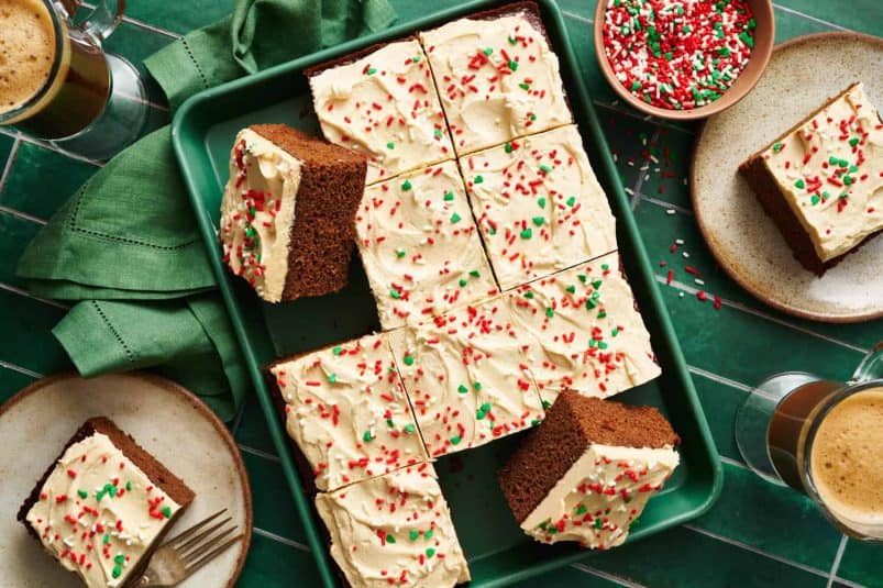 Overhead view of a rectangular gingerbread cake with frosting on top and red and green sprinkles on a green background with a green napkin.