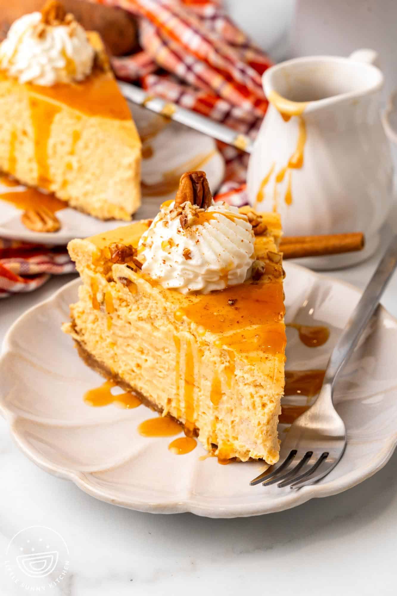 A dessert plate holding a metal fork and a slice of sweet potato cheesecake with whipped cream. in the background is a small pitcher of caramel sauce and another slice of cheesecake.