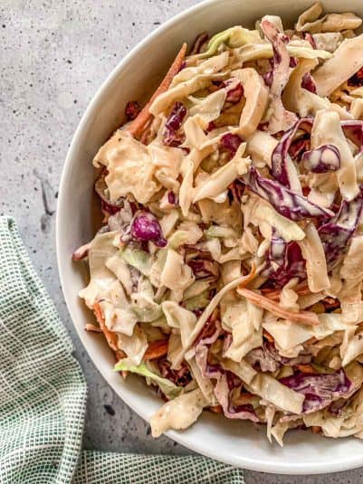 A bowl of creamy coleslaw featuring shredded cabbage in green and purple hues, mixed with carrots. The coleslaw is placed on a speckled gray surface beside a green and white patterned cloth.