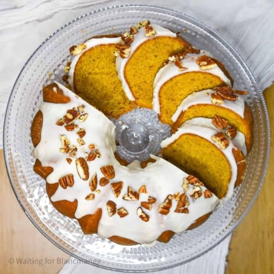 Overhead picture of pumpkin pound cake, topped with vanilla glaze and chopped pecans. Half unsliced and half sliced and laid out on the platter.