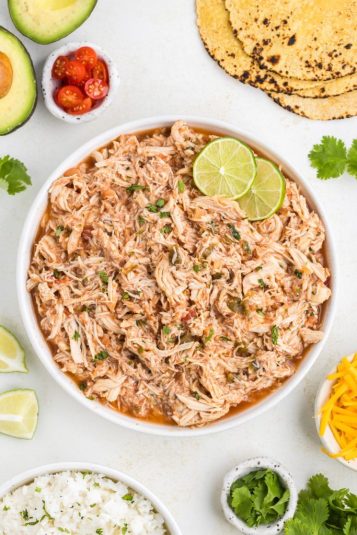 Bowl of slow cooker salsa chicken surrounded by tortillas, rice and condiments.