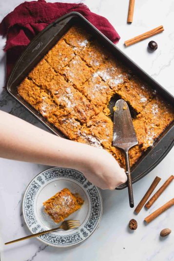 A person slices a baked dessert, reminiscent of a carrot casserole, in a rectangular pan on a marble surface. The orange treat is dusted with sugar. A plated piece with a fork sits on the left, surrounded by cinnamon sticks and a red cloth.
