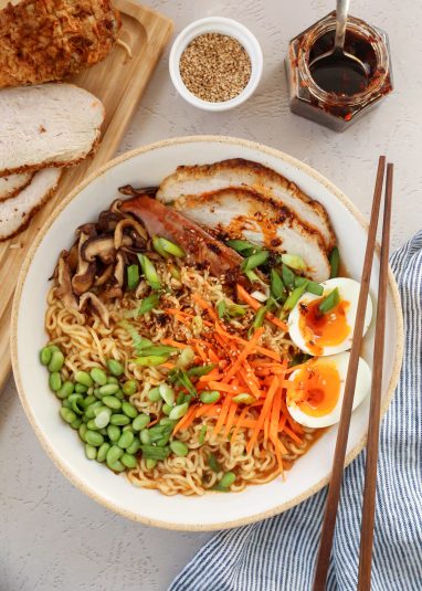 Overhead image of a bowl of leftover turkey ramen, with thick slices of roasted turkey resting inside a ramen bowl topped with carrots, edamame, soft boiled 7 minute eggs, green onions, and mushrooms and a drizzle of chili oil on top.