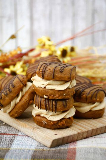 A pile of pumpkin whoopie pies on a wooden cutting board sitting on a checkered cloth.