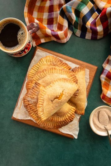 Pumpkin Cheesecake Empanadas are stacked in a pile on a wooden board next to a mug of coffee and a plaid kitchen towel.