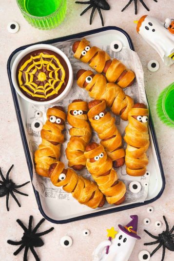 Golden Air Fryer Mummy Hot Dogs on a platter with small bowls of ketchup and mustard spider web dip for Halloween.