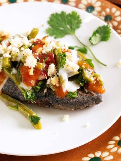 Closeup view of a bitten into blue corn tlacoyo that's filled with black beans and topped with ensalada de nopales, sour cream, salsa, and vegan cotija cheese.