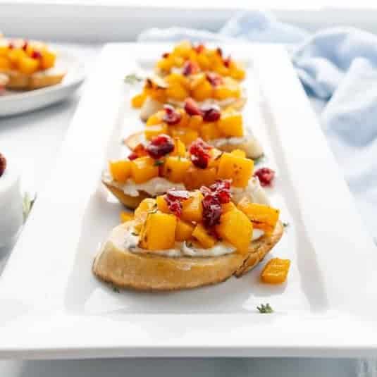 A white rectangular plate features toasted slices of bread topped with ricotta cheese, diced squash, and cranberries.