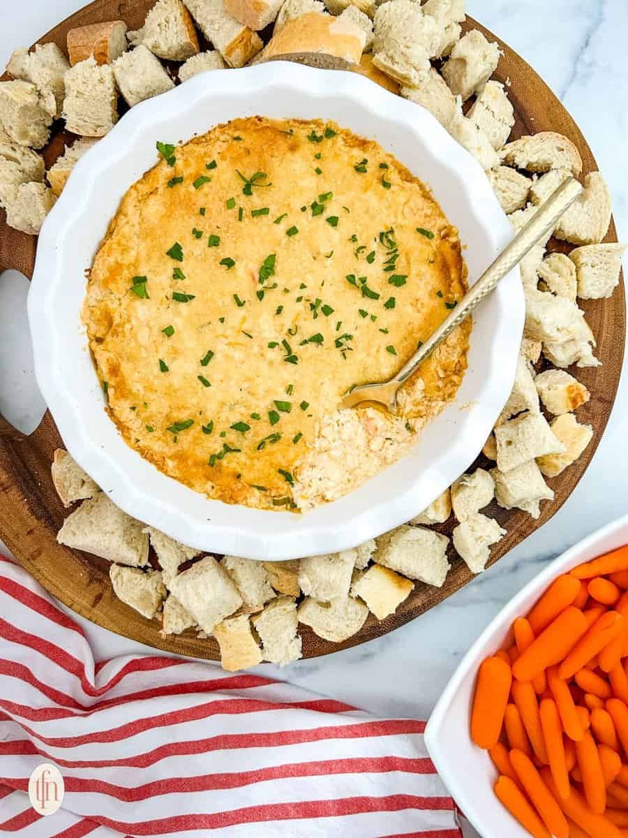 A bowl of cajun crab dip with shrimp with croutons served on a wooden platter with a bowl of carrots.