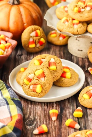 A plate with freshly baked candy corn Halloween cookies sits on a wooden table. Additional candy corn and cookies are scattered around, and a pumpkin can be seen in the background.