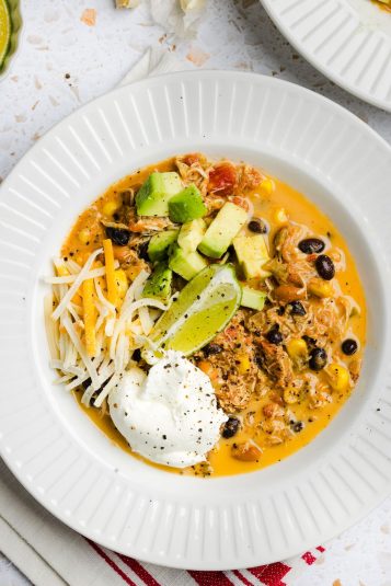 Overhead photo of creamy red chicken chili in a white bowl, garnished with avocado and greek yogurt.