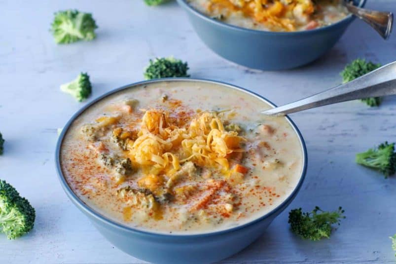 Broccoli cheese soup in a blue bowl with a spoon, with broccoli scattered around.
