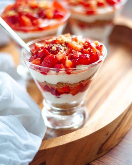 Close up of a stemless martini glass filled with this deconstructed cheesecake with strawberries. The glass sits has a spoon in it and sits on a wooden tray with a white linen and additional glasses filled with strawberry no bake cheesecake.