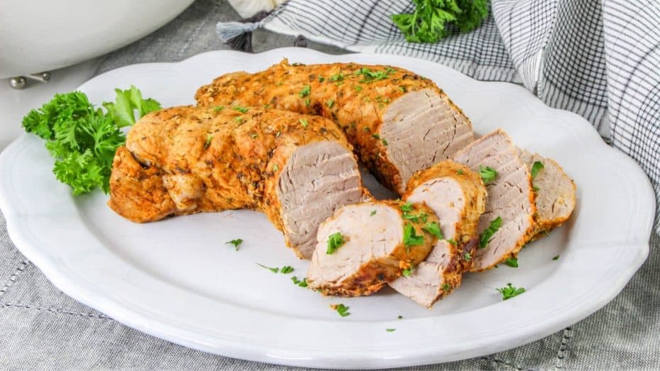 Sliced roasted pork tenderloin garnished with parsley on a white plate, with a striped cloth in the background.