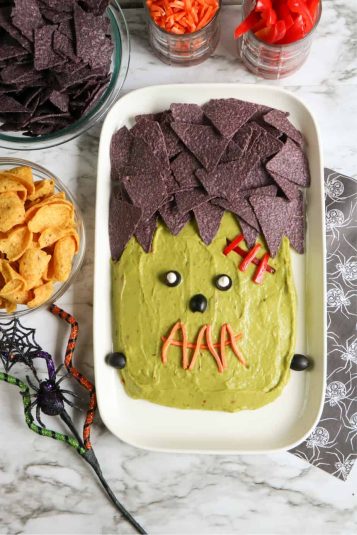 Plater of guacamole decorated to look like Frankenstein with jars of chips and veggies to dip in bowls.