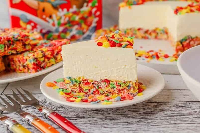Slice of Fruity Pebbles cheesecake on a plate, with rest of cake and Rice Krispie Treats with Fruity Pebbles in the background.