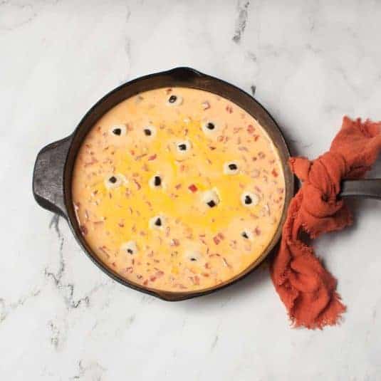 A cast iron skillet with Halloween cheese dip in it. There is an orange linen tied to the handle.