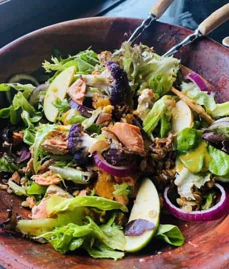 A wooden bowl with salad tongs surrounded by a green salad filled with pears, salmon and onions.
