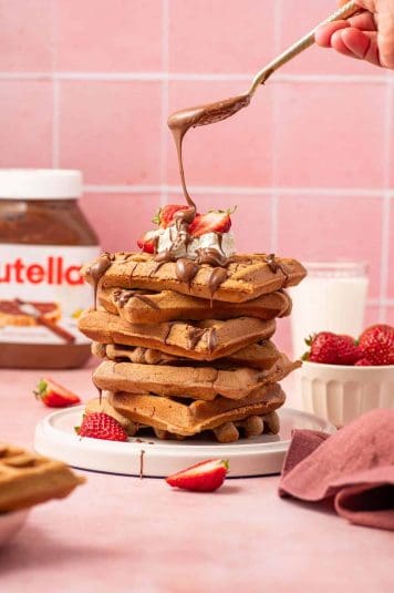 A stack of warm Nutella waffles served topped with whipped cream, fresh strawberries, and a rich chocolate-hazelnut drizzle, creating a decadent and mouthwatering brunch treat.