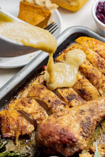 Gravy being poured over sliced roasted turkey breast on a sheet pan, with cranberry sauce and pumpkin pie visible in the background.