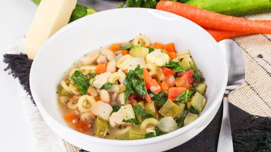 A white bowl of vegetable soup with pasta rings, carrots, zucchini, and greens, placed on a table with a spoon and carrot in the background.