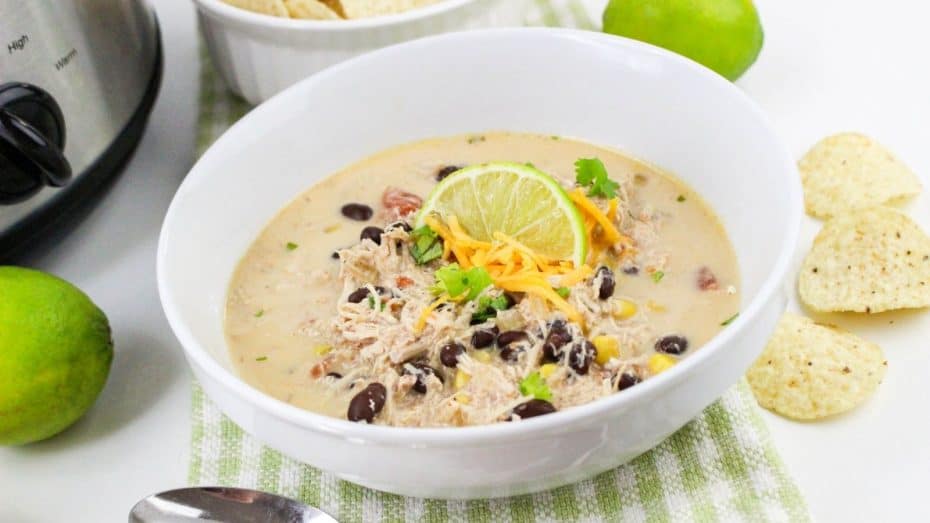 Bowl of creamy chicken soup with shredded chicken, black beans, corn, a lime slice, and cheese on top. Lime, tortilla chips, a spoon, and a slow cooker in the background.