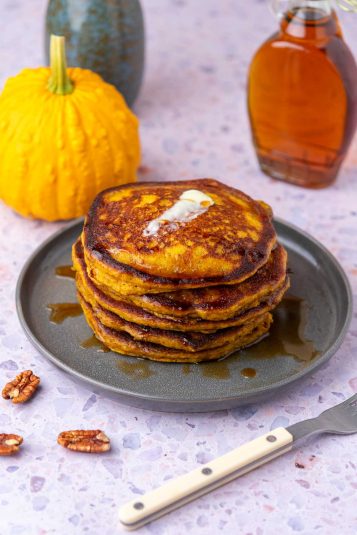 Stack of sourdough pumpkin pancakes on a plate with pecans, butter, and maple syrup.