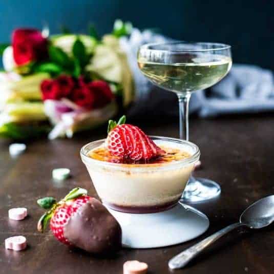A serving of vanilla french toast next to a serving plate and bowls of candied pecans, blueberries, powdered sugar, and a banana.
