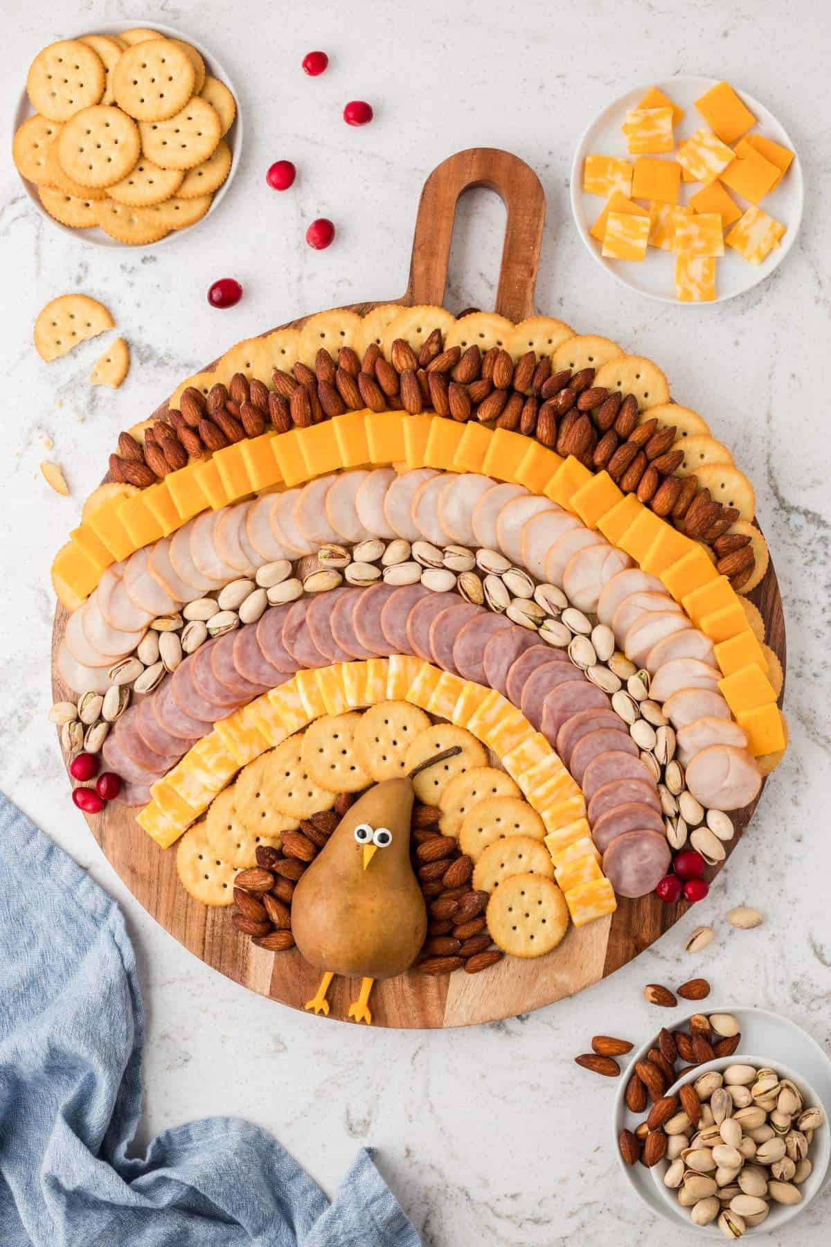 A Thanksgiving charcuterie board arranged in the shape of a turkey, featuring slices of cheese, meat, nuts, crackers, and a pear as the turkey's body. Small dishes with extra snacks are placed around the board.