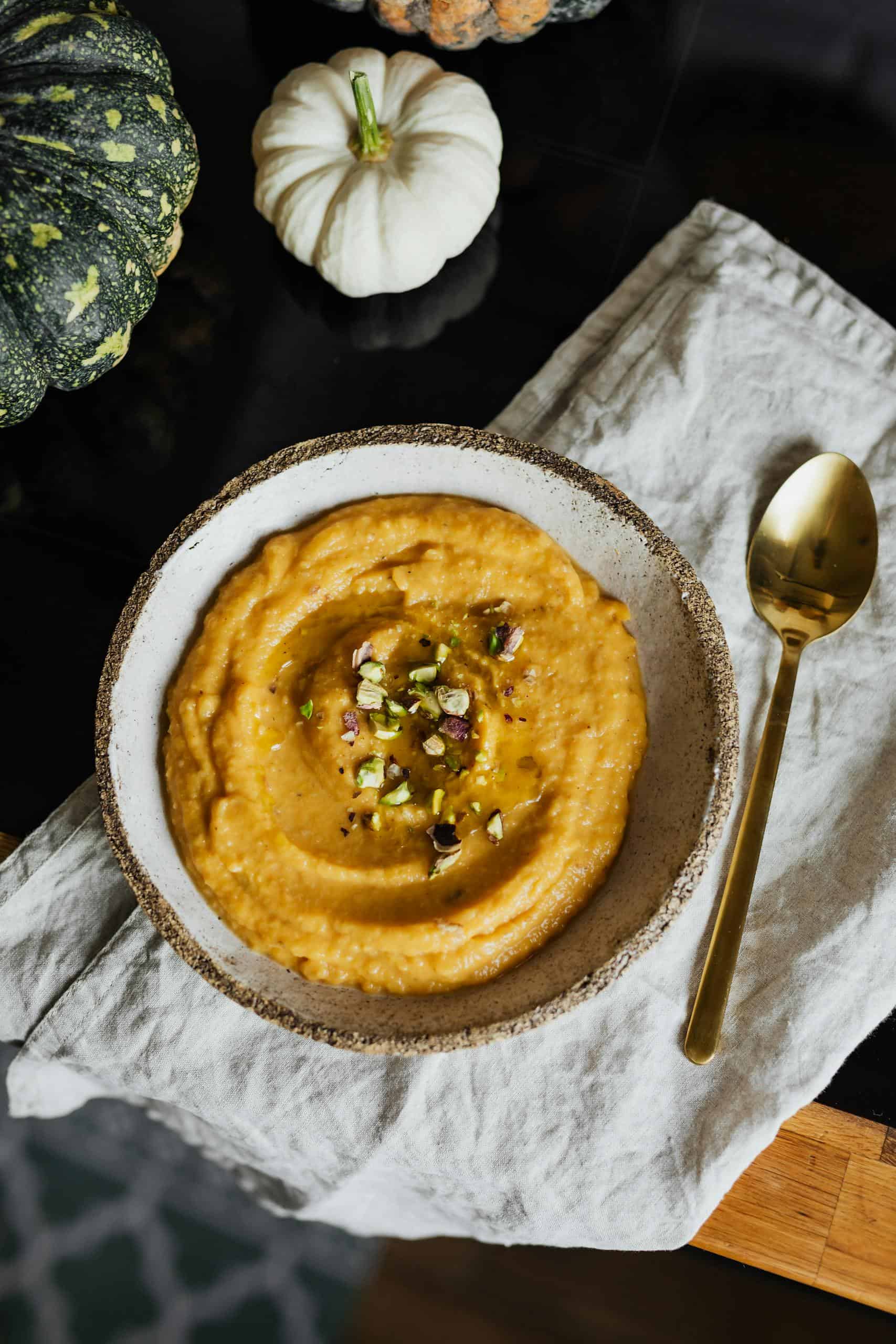 A creative flatlay of pumpkin hummus garnished with pistachios and a gold spoon.
