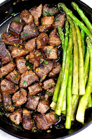 Steak bites drenched in garlic butter sauce, alongside some asparagus.