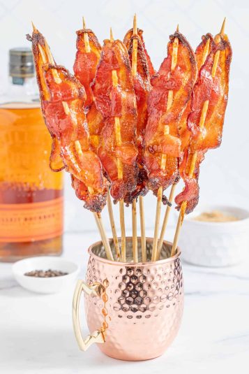Several candied bacon skewers standing upright in a copper mug on a white counter top with a bottle of bourbon and a white ramekin of brown sugar.