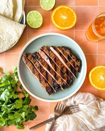 Half of a carne asada breakfast burrito is held over a blue plate surrounded by limes, orange juice, coffee, and a white linen.