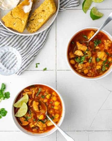 Two bowls of southwest chicken soup on a white table with a plate of cornbread.