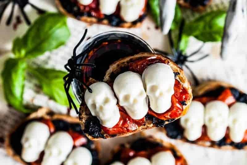 Toasted bread topped with cherry tomatoes and three white skull-shaped cheese pieces, this Halloween bruschetta appetizer is served with spooky Halloween decor.