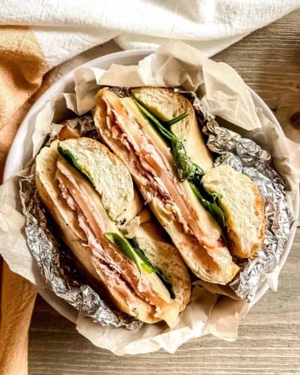 Baked ham and cheese bagel sandwich wrapped in foil and parchment paper, in a bowl.