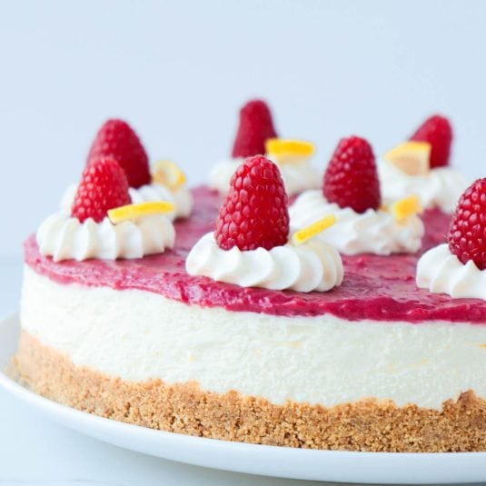 Cheesecake topped with whipped cream, fresh raspberries, and wedges of lemon.