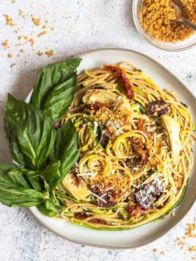 Mediterranean zucchini spaghetti on a plate next to a bowl of toasted breadcrumbs with a spoon.