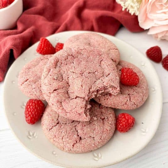 Raspberry Cookies stacked on a white plate with fresh raspberries, one cookie with a bite missing.